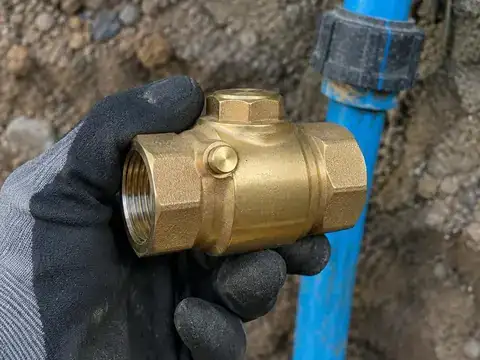 Gloved hand holding a new brass check valve near a blue well water line